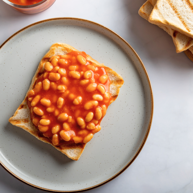 Toast with baked beans on a white plate, with additional toast and a bowl in the background.